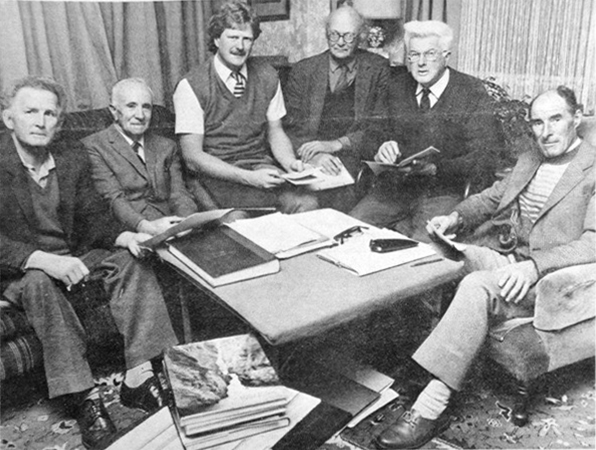 A planning meeting with 6 elderly men in dated clothing sitting around a table with reference books etc.